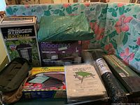 Full view of all items in the lot arranged on a floral outdoor cushion including the bug killer in box, path lights box, greenhouse kit box, picnic table cover, lounge chair pad, vinyl-coated wire roll, tarp, and garden tool bag.