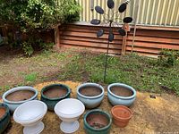 Wide arrangement of six plant pots including large ceramic, plastic urns, and terra cotta, with metal windmill behind on lawn.