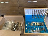 Two boxes: wooden flatware storage box with appear 12 knives in lid and assorted flatware pieces in tray; adjacent cardboard box containing loose forks and spoons.