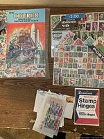 Photo of The Courier stamp album, loose stamps, and Dennison pre-folded stamp hinges on wooden surface.