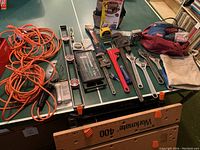 Photo showing assorted hand tools laid out on green surface including several levels, large wrenches, adjustable wrenches, pipe wrenches, Ryobi power sander in bag, extension cords, and a small tool bag.