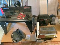 Shows the router table with its metal surface, manual, router bits and parts, alongside the Craftsman bench grinder mounted on wooden base and a box of sharpening stones.