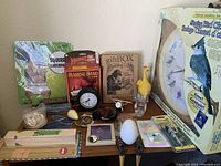 Overview of all items in the lot on a table including singing bird clock, harmonicas, rubber chicken, novelty items