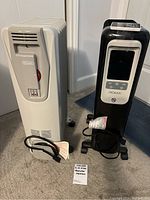 Two oil-filled radiator heaters side by side, black Noma and grey DeLonghi heater, both on carpeted floor with cords neatly coiled