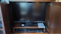 Front view of the Sharp LC-42D64U 42 inch LCD TV inside a wooden cabinet, showing the screen, remote, and manual on the shelf below.