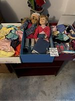 Two dolls inside a box surrounded by doll clothes and accessories on a table.