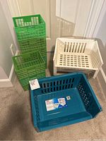 Four assorted plastic bins: three green crate style bins stacked, one white jumbo stacking basket, and one blue jumbo stacking basket on carpet floor.