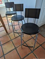 Front and side view of two aluminum frame bar stools with black plastic seats and backs placed on tiled floor near a window and door.
