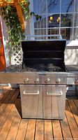 Front view of stainless steel KitchenAid propane barbecue grill with four control knobs, open lid showing grill grate inside, and two-door lower cabinet storage.