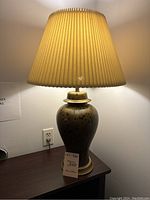 Full view of the lit table lamp showing brown and black splattered base with gold accents and the cream pleated shade.