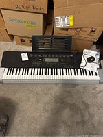 Casio WK-240 electric keyboard front view with music stand attached and power adapter cable placed on top. Background includes cardboard boxes.