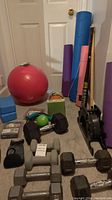 Photo showing two pairs of gray dumbbells, several black dumbbells, blue yoga blocks, colorful yoga mats (blue, purple, pink), green exercise ball, and a black rowing machine.