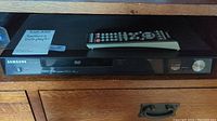 Samsung black DVD player with remote on top, front panel showing power button and disc tray.