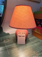 Full view of the small lamp lit on a table showing the pink shade and base.