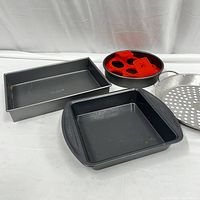 View of all four items: rectangular and square black non-stick pans, round pan with red silicone egg molds, and metal steamer insert basket.