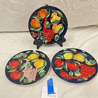 Photo showing the set of three round pottery dishes each hand painted with fruit motifs on a dark blue background, displayed on a white surface with one upright on a black stand.