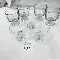 Six clear glass footed bowls arranged in two rows on white fabric background. Four larger individual trifle/fruit bowls and two smaller dessert glasses.