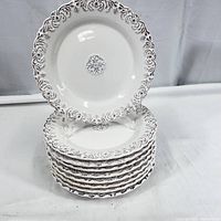 Stack of nine white ceramic dinner plates with embossed floral and scroll ornate border in brown.