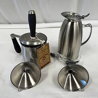 Photo showing all four items: basting pot with handles and lid, tall pouring jug, two funnel shaped items, arranged on white backdrop.