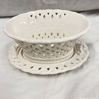 Front view of white ceramic oval pierced scalloped bowl sitting on matching under plate with grape cluster handles.