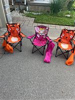 Three kids camping chairs set outside on pavement: two tiger-themed orange chairs with tiger tails and one pink princess chair.
