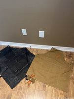 Both aprons laid out side by side on a floor showing the black work apron and the neutral tan canvas kitchen apron with leather straps.