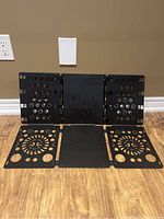 Black plastic folding board unfolded on wooden floor showing six panels with ventilation holes