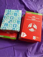 Four packs of copy paper stacked on a purple background, showing red Staples brand packs and a blue and white HOTS cover pack