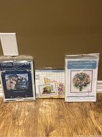 Three cross stitch kits sealed in plastic packaging, showing front designs: dragon scene, flower bouquet, colorful detailed pattern.
