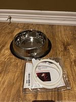 Stainless steel dog bowl with black rubber base placed on wooden floor next to packaged LED dog collar.