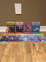 Photo showing the full set of 9 R.L. Stine Goosebumps books arranged in two rows on a wooden floor against a beige wall.