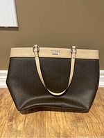 Front view of Guess brown and cream tote purse on wooden floor against tan wall.