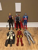 Six Marvel action figures arranged in two rows on floor: Hawkeye, Thor, Captain America standing in back row; Venom, Iron Man, and silver-armored figure lying down in front row.
