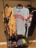Men's clothing items on hangers displayed on a clothing rack, featuring a beige and black tie-dye sweatshirt, a Houston Oilers t-shirt, a Georgia Bulldogs t-shirt, and other folded graphic t-shirts.