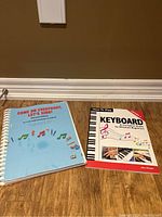 Two music books placed side by side on a wooden floor, one with a light blue cover titled 'Come On Everybody, Let's Sing!' and the other titled 'How to Play Keyboard' for beginners.