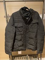 Front view of black Oxford Tech winter coat showing padded quilting on arms, pockets, and front area, hanging on hanger.