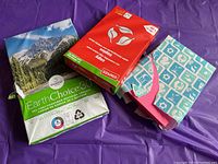 Lot of paper packs and reams on purple surface including Domtar EarthChoice ream, red Staples copy paper pack, neon pink cardstock, and patterned paper pack.