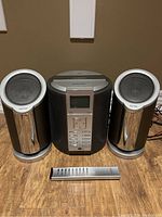 Central audio unit flanked by two speakers and remote control in front, placed on wooden floor.