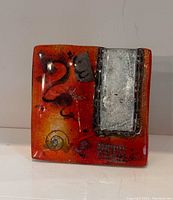 Square fused glass bud vase with red-orange background, silver rectangular panel, and black abstract designs.