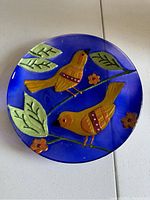 Full view of the round blue decorative plate with two yellow-orange birds, green leaves, and orange flowers in raised relief.