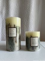 Front view of two new botanical candles of different heights on a white surface with a textured backdrop.