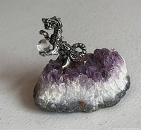 Close-up image of a small pewter dragon statue holding a faceted crystal ball standing on an amethyst crystal cluster with purple and white points.
