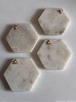 Four white marble hexagon coasters arranged showing etched honeycomb pattern and small gold bee figurine on top corner of each coaster.