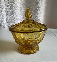 Amber-colored Indian glass compote with lid and finial detail on a pedestal base photographed against a white surface and backdrop.