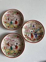 Photo of three Japanese miniature plates: two with matching design showing three women in traditional attire surrounded by flowers, one with different scene including people on a bridge and a building. One plate has a visible crack.