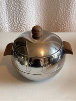 Front view of stainless steel round server pot with wooden handles and lid knob, showing the smooth reflective surface and embossed penguin decoration.