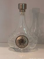 Front view of clear glass decanter with silver-plated medallion and stopper, placed on a white surface.