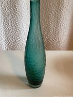 Front view of the tall turquoise textured glass vase placed on a white surface against a textured white wall background.