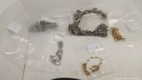 Full view of five jewelry pieces including silver and gold-toned chains and bracelets in plastic bags on white background