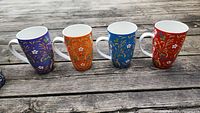 Four porcelain mugs with unique floral patterns in purple, orange, blue and red arranged in a row on a wooden surface.